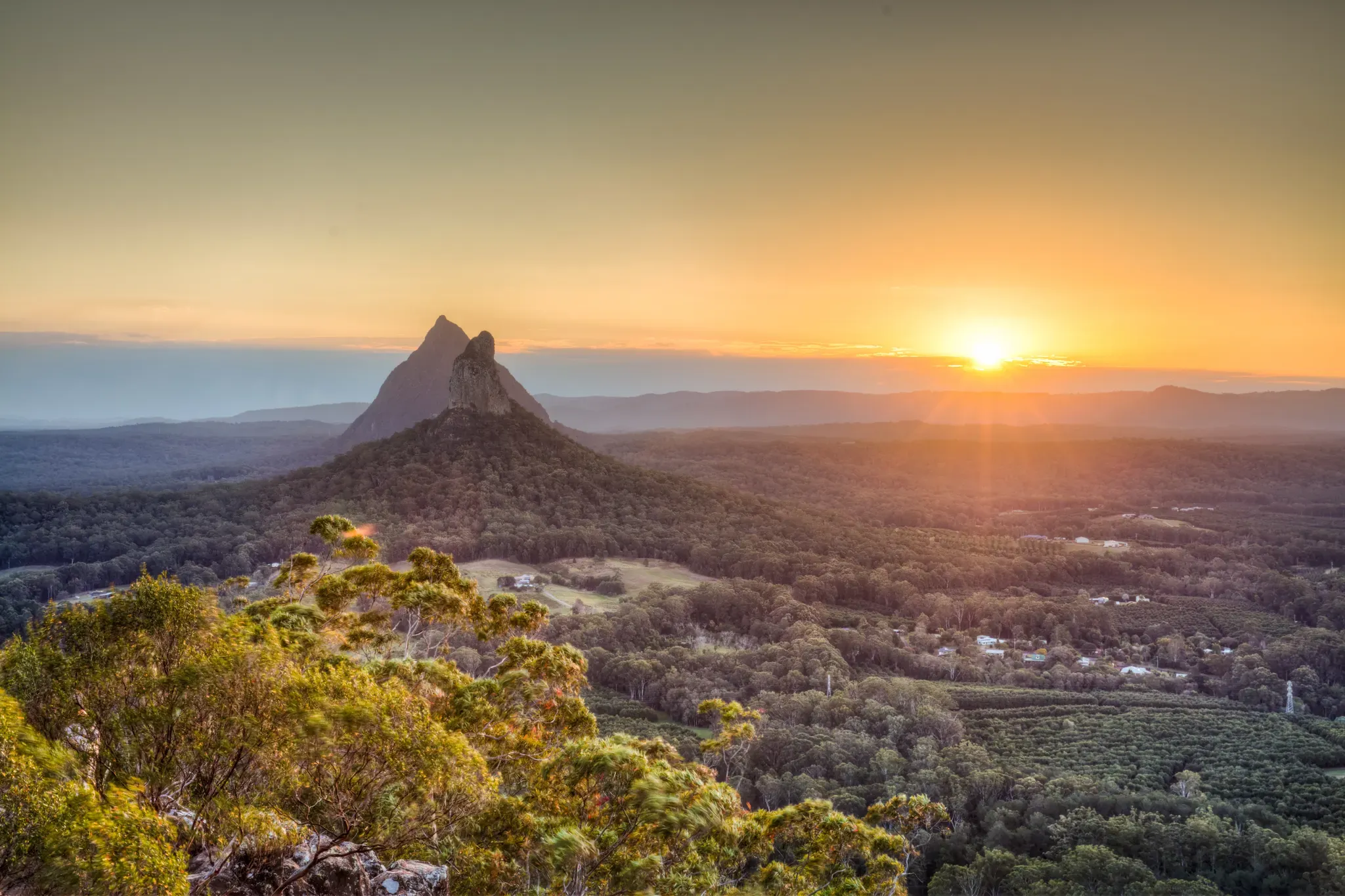 A scenic view of lush green forest, scattered houses, and a distinct mountain peak at sunrise, with the sun casting a golden glow over the landscape and distant hills under a clear sky.