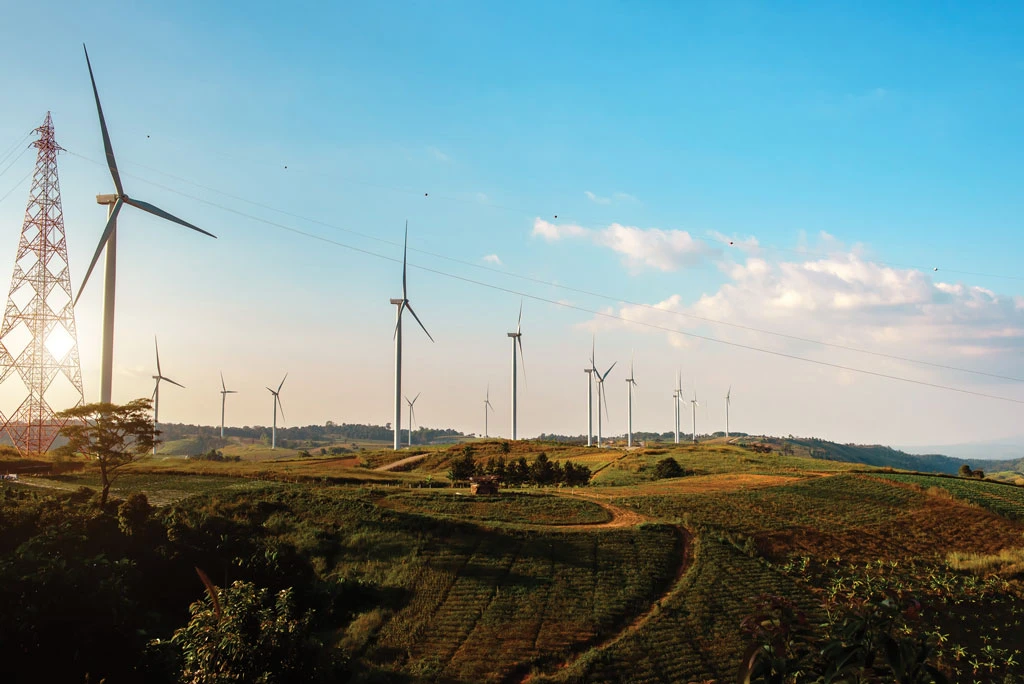 A landscape with multiple wind turbines and a tall electricity pylon standing on grassy hills under a blue sky with scattered clouds, captured during daylight.