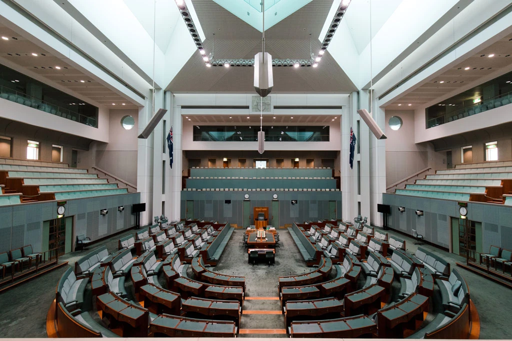 A wide view of an empty parliamentary chamber with curved rows of desks and chairs, high ceilings, Australian flags, and a central speaker’s podium at the front.