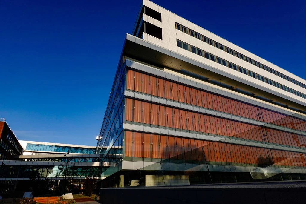 A modern multi-story building with glass and reddish-brown paneling, set against a clear blue sky, reflecting its surroundings and connected by a glass walkway.