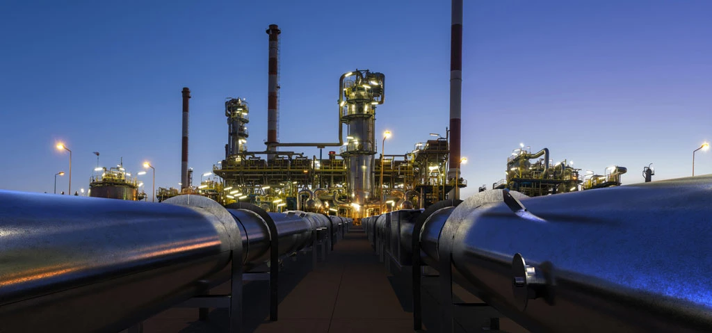 A large oil refinery at dusk, with illuminated towers and pipes stretching into the foreground. The industrial scene is set against a clear evening sky.
