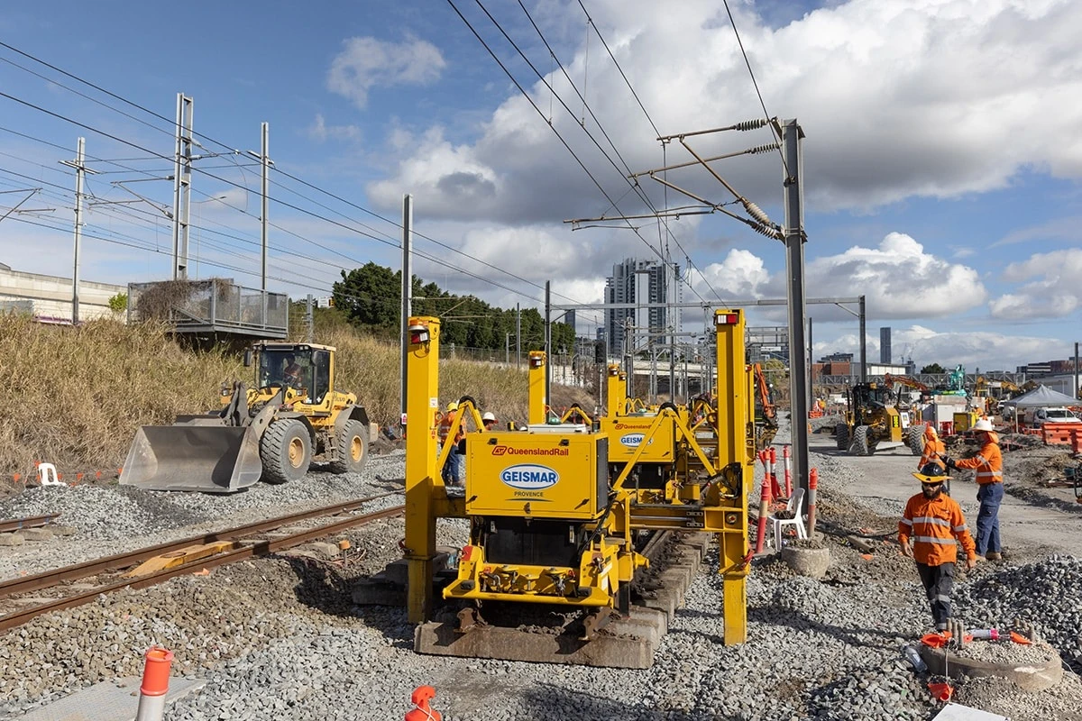 Workers in orange safety gear operate heavy machinery on a railway construction site, with tracks being installed and gravel spread. Equipment and power lines are visible under a partly cloudy sky near city buildings.
