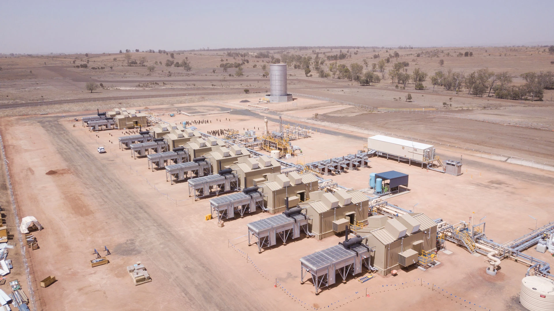 Aerial view of a large industrial facility with multiple beige machinery units arranged in rows on a cleared, dusty landscape, surrounded by dry, sparsely vegetated terrain under a clear sky.