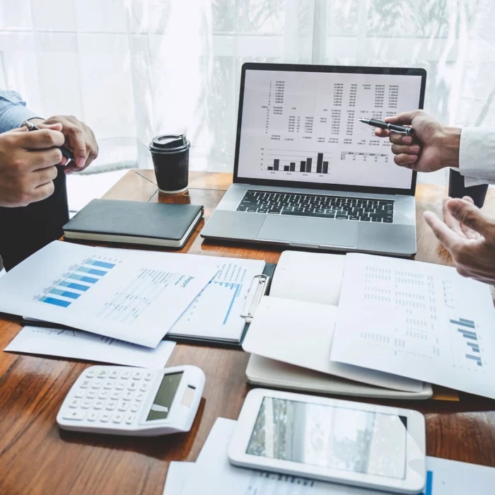 Two people discuss financial charts and graphs at a desk with documents, a laptop displaying analytics, a calculator, a tablet, notebooks, and a coffee cup. One person gestures with a pen.