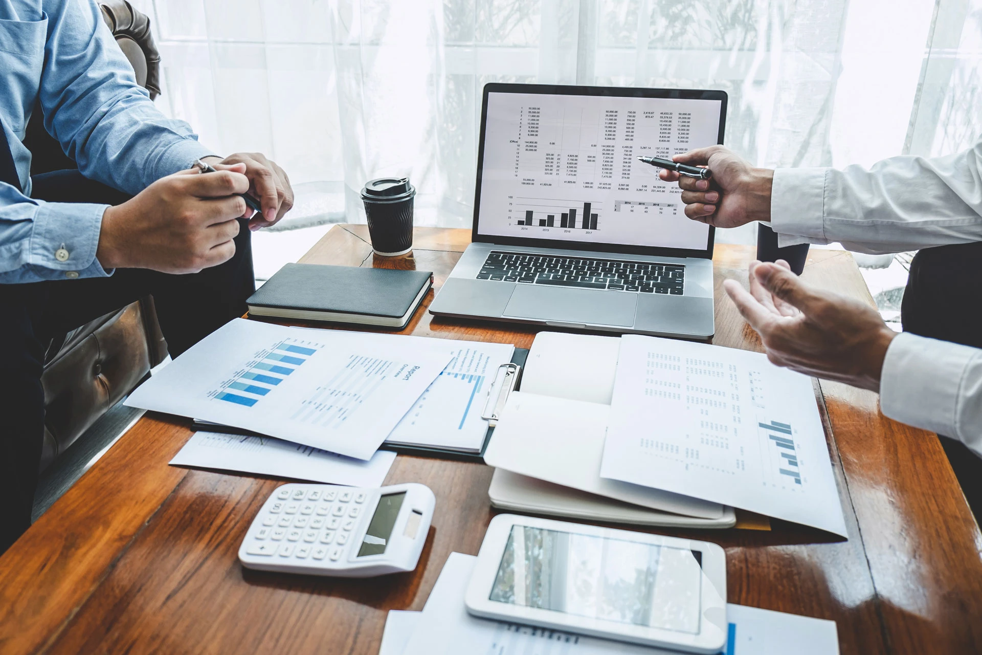 Two people discuss financial charts and graphs at a desk with documents, a laptop displaying analytics, a calculator, a tablet, notebooks, and a coffee cup. One person gestures with a pen.