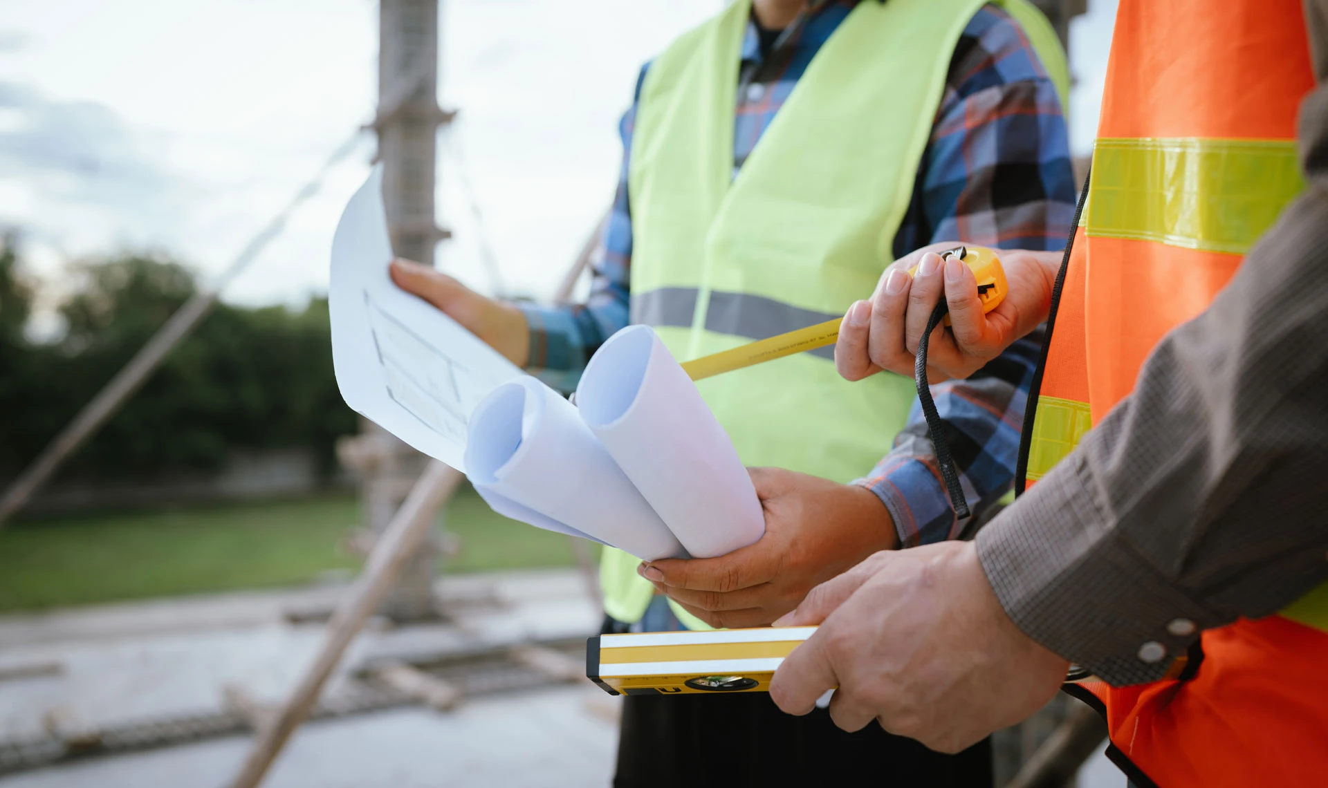 Two construction workers wearing safety vests are holding blueprints, a tape measure, and a spirit level, discussing plans at an outdoor building site. Only their upper bodies and hands are visible.