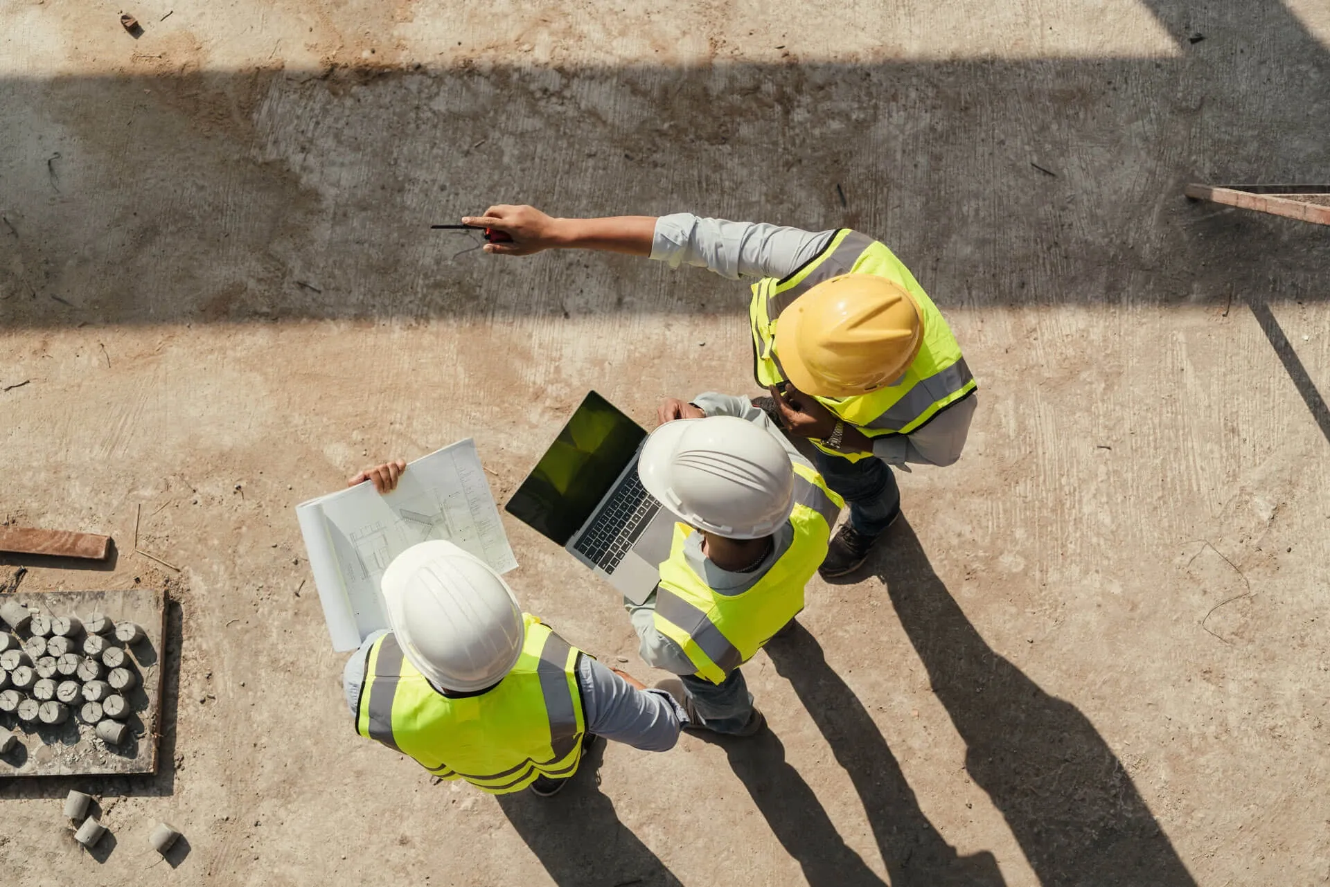 Three construction workers in safety vests and helmets stand on a site, looking at a laptop and blueprints as one points ahead, likely discussing project plans. The scene is viewed from above.