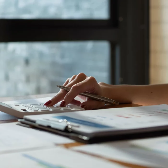 A person sits at a desk with documents, charts, and a calculator, pressing buttons while holding a pen. Glasses and a small potted plant are nearby, with sunlight coming through a window in the background.