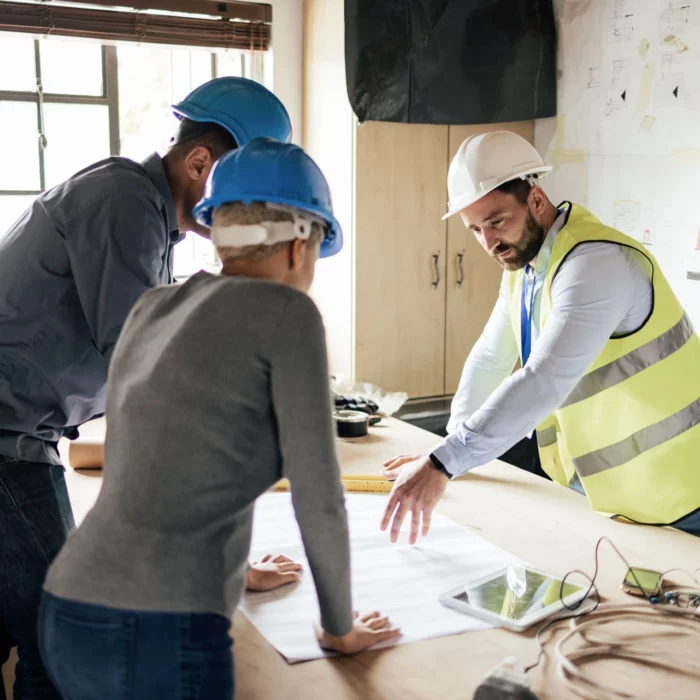 Three people wearing safety helmets and a reflective vest discuss plans at a table with blueprints, a tablet, and construction documents in a bright office with project drawings on the wall.