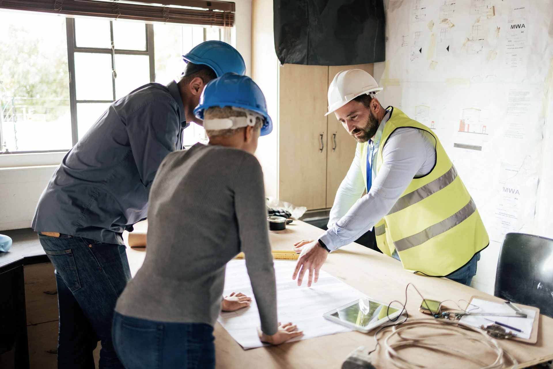 Three people wearing safety helmets and a reflective vest discuss plans at a table with blueprints, a tablet, and construction documents in a bright office with project drawings on the wall.